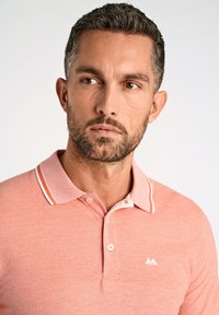 T-shirt med kort ärm i korall med en krage som har vita detaljer och en logotyp på vänster sida. Mjuk textur och subtilt mönster.