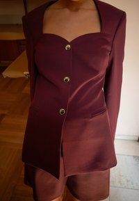 Burgundin värinen strukturoidu pakojakku, jossa on sydänkaula-aukko, kultaiset napit ja sivutaskut, yhdistettynä samasta sileästä kankaasta valmistettuihin shortsihousuihin.