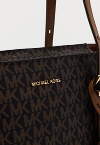 Schwarze Stoff-Shopper-Tasche mit einem wiederholten dunkelbraunen "MK"-Muster. Verfügt über ein goldenes "MICHAEL KORS"-Logo und braune Lederakzente an den Tragegriffen.