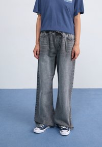 Vide ben, grå denimbukser med en falmet tekstur, som har en elastisk talje og snor, parret med blå kanvas sneakers.
