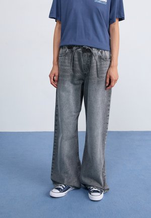 Wide-leg, gray denim pants with a faded texture, featuring an elastic waistband and drawstring, paired with blue canvas sneakers.