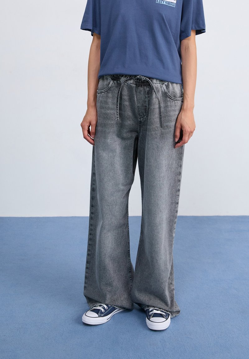 Vide ben, grå denimbukser med en falmet tekstur, som har en elastisk talje og snor, parret med blå kanvas sneakers.