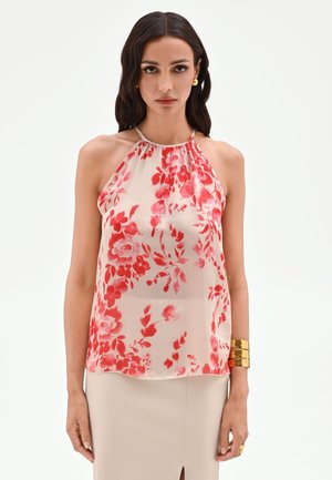 Femme portant un chemisier sans manches à col halter avec imprimé floral rouge, jupe beige fendue à l'avant, bracelet manchette doré et boucles d'oreilles dorées.