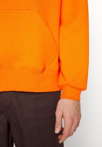 Sudadera naranja con un bolsillo tipo canguro y puños acanalados, combinada con pantalones marrón oscuro. La tela parece suave, con una textura lisa.