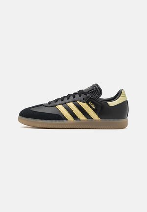 Zapatilla Adidas Samba negra y dorada con suela de goma, con tres rayas doradas y cordones negros, mostrada sobre un fondo blanco liso.