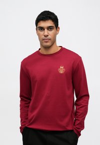 Bordowa koszulka z długim rękawem wykonana z miękkiej tkaniny, z złotym haftowanym wzorem na piersi i okrągłym dekoltem. Dopasowany krój.