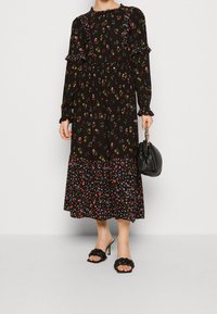 Abito nero a fiori con maniche lunghe e dettagli a volants, caratterizzato da una gonna fluida e a strati. Abbinato a una borsa nera e sandali con tacco.