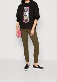 Svart sweatshirt med en färgglad Minnie Mouse-grafik, olivgröna smala byxor och svarta slip-on-skor med vita detaljer. Rosa handväska.