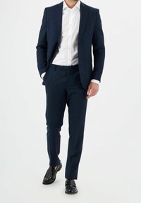 Tmavě modrý oblek s proužky, bílá košile a černé boty. Oblek má na míru šitý střih a výrazné klopy.