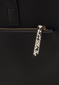 Bolso de cuero negro con una cremallera dorada y un tirador de cremallera distintivo con patrón de serpiente y acentos en negro y blanco.