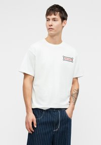 T-shirt en coton blanc à manches courtes et col rond, avec un logo "VOLCOM" imprimé en rouge et gris sur la poitrine gauche.
