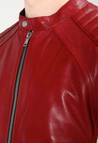 Rode leren jas met een gladde textuur, voorzien van een hoge kraag, ritssluiting en gequiltte schouderaccenten. De rits heeft een metallic afwerking.