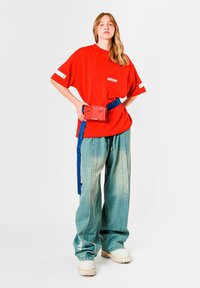 Camiseta roja de gran tamaño con un logo blanco, combinada con pantalones anchos de mezclilla azul claro y una riñonera roja. Zapatos beige con suela gruesa.