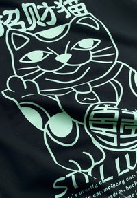 Graphique stylisé de chat porte-bonheur avec la patte levée, clochette au collier, et caractères chinois au-dessus, imprimé sur un tissu foncé avec un texte anglais partiel en dessous.
