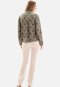 Chemise à manches longues, à motifs dans des nuances de bleu et doré, en tissu léger, associée à un pantalon beige ajusté et des chaussures beiges.