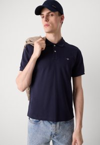 Marinblå pikétröja med korta ärmar, treknappslåsstängning och logga. Ljusbeigefärgad tröja draperad över axeln, som bärs med ljus denimjeans.