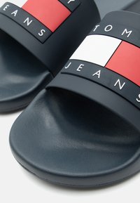 Tengerészkék csúszós szandál piros, fehér és tengerészkék márkázott pánttal, kiemelkedő "Tommy Jeans" felirattal.