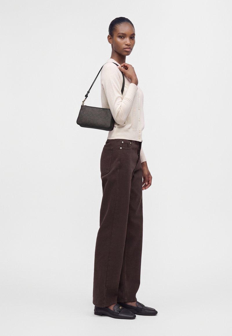 Femme debout de profil portant un cardigan beige clair, un pantalon large marron foncé, des mocassins noirs et portant un petit sac à épaule noir.