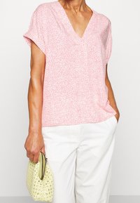 Helles rosa Kurzarmbluse mit V-Ausschnitt; zeichnet sich durch ein dezentes gesprenkeltes Muster aus. Kombiniert mit weißen Hosen und einer geflochtenen gelben Handtasche.