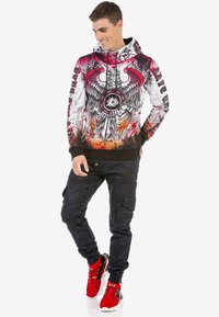 Kapuzensweatshirt mit rotem und schwarzem Grafikdesign, das einen Adler, Flammen und einen Schädel zeigt. Kombiniert mit dunklen Tarnhosen und roten Turnschuhen.