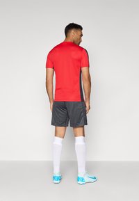 Punane sportlik särk lühikeste varrukate ja mustade detailidega, kombineeritud tumehallide triibuliste lühikesed püksid ja valged sokid, seljas sinised tossud.