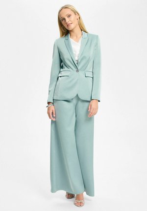Femme blonde portant un blazer bleu clair cintré et un pantalon à jambes larges avec un haut blanc et des talons ouverts, debout sur fond blanc.