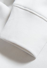 Puño de sudadera blanca con un tejido texturizado, acabado suave y un detalle de costura limpio. El tejido parece ligero y transpirable.