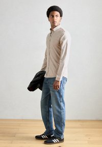 Ανοιχτό μπεζ πουκάμισο με υφή, συνδυασμένο με χαλαρά μπλε τζιν και μαύρα sneakers με λευκές ρίγες, κρατώντας μια μαύρη ζακέτα.