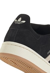 Baskets en daim noir avec une semelle blanche texturée, présentant un logo accent blanc au talon et des détails cousus le long du bord.