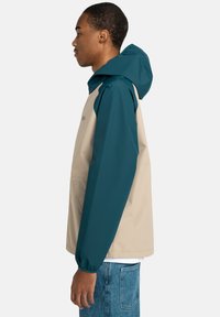 Kleurblokjas met een beige lichaam en teal mouwen, gemaakt van lichtgewicht materiaal met een capuchon en een kangoeroezak.