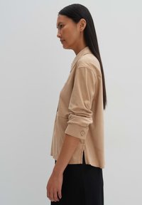 Beige overhemd met lange mouwen en een gekraagde halslijn, opgerolde manchetten, zijsplitten en een gladde, zachte textuur. Eenvoudig, minimalistisch ontwerp.