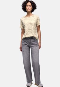 Nainen, jolla on kiharat hiukset, beige leopardikuvioinen lyhythihainen paita, harmaat suorat farkut ja harmaat lenkkarit, seisoo yksiväristä taustaa vasten.