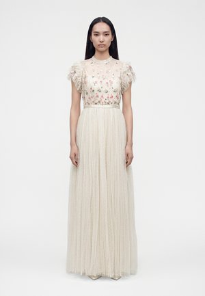 Femme portant une longue robe beige en tissu transparent à pois, broderie florale sur le corsage et manches courtes à volants, debout devant un fond blanc.