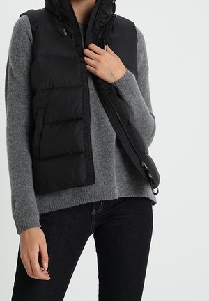 Person trägt einen grauen Pullover und eine schwarze Daunenweste, hält den Reißverschluss der Weste mit einer Hand und ist in schwarzen Hosen vor einem einfarbigen Hintergrund gekleidet.