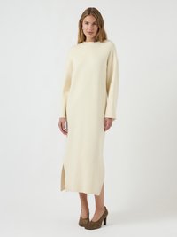 Abito lungo color crema realizzato in morbido tessuto a maglia, con collo alto, maniche lunghe, spacco laterale e una vestibilità rilassata. Abbinato a tacchi marroni.