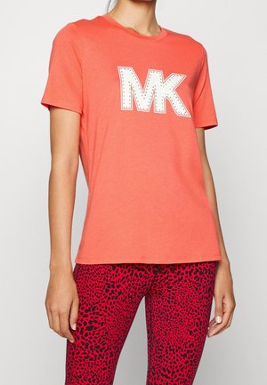 Nainen, jolla on päällään korallinsävyinen lyhythihainen t-paita, jossa valkoinen "MK"-logo, sekä punamustat pilkulliset leggingsit, seisoo yksinkertaisen taustan edessä.