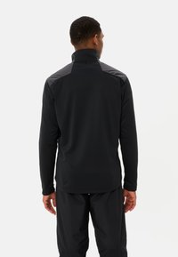 Man in een zwart en donkergrijs lang-sleeve jasje en zwarte broek, weergegeven vanaf de achterkant tegen een effen lichte achtergrond.