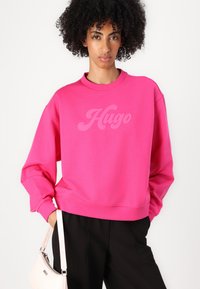 Felpa rosa con un grande e discreto logo "Hugo". Collo e polsini a costine. Indossata con pantaloni neri e una borsa leggera.