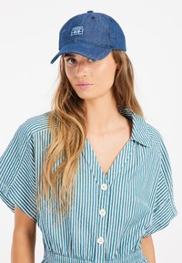 Denim-basebollkeps med broderad "PROTEST"-logotyp, böjd skärm och sexpanelig konstruktion, med en texturerad väv i mörkblått.