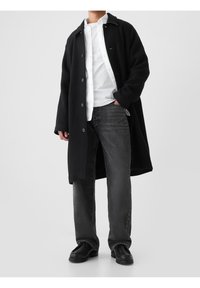 Manteau en laine noire avec fermeture par boutons sur une chemise blanche à boutons, associé à un jean gris foncé et des chaussures noires. Design simple, sans ornements.