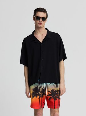 Uomo con camicia nera a maniche corte con bottoni, occhiali da sole e pantaloncini rossi con stampa di palme nere, in piedi davanti a uno sfondo semplice.