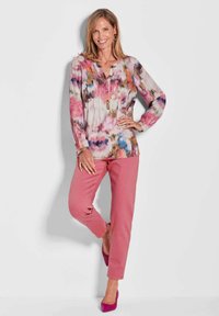 Mehrfarbige Bluse mit floralem Muster und Knöpfen am Ausschnitt; kombiniert mit einfarbigen rosa Hosen und rosa High Heels. Glatte Stofftextur.