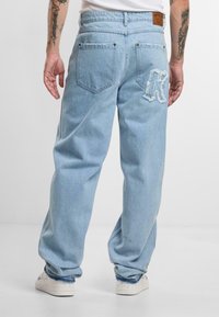 Lyseblå denimjeans med en avslappet passform, med slitte detaljer og en brodert logo på baklommen. Brukt med hvite sneakers.