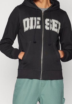 Person wearing black zip-up hoodie with "DIESEL" printed in large white letters across chest and green pants, hands in pockets.