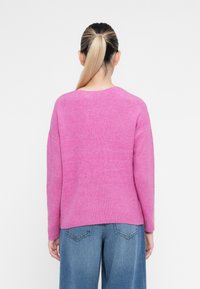 Sweater rosa com relevo, com decote redondo e mangas longas, combinado com jeans azuis de perna larga, visto pela parte de trás.