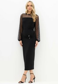 Czarna sukienka z przezroczystymi długimi rękawami w kropki, dopasowanym dekoltem, marszczoną talią i metalowym akcentem. Gładki materiał, długość do kolan.