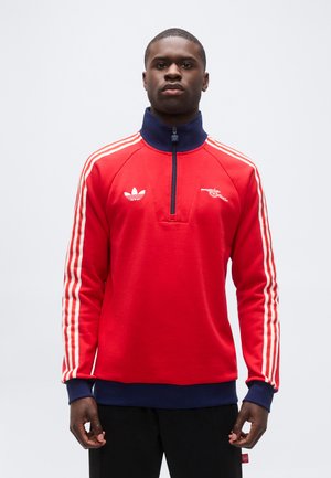 Mann trägt rotes Sweatshirt mit Reißverschluss, weißen Streifen an den Ärmeln und Sportlogos, steht vor hellem, einfarbigem Hintergrund.