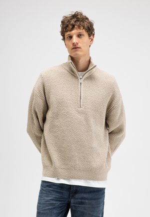 Young man with curly hair wearing a beige knitted half-zip sweater over a white shirt and blue jeans, standing against a plain background.