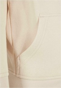 Nahaufnahme einer beigen Stofftasche auf einem Sweatshirt mit sichtbaren Nähten und einem gerippten Bündchen am unteren Rand.