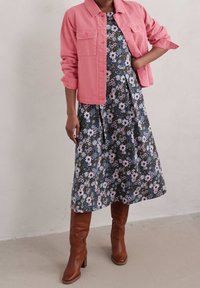 Světle růžová denimová bunda, květinové tmavě modré šaty s fialovými a bílými květy, hnědé kozačky na vysokém podpatku po kolena, šaty střední délky, ležérní střih.